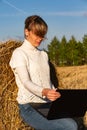 Pretty girl on a background of blue sky in a field near a haystack in a white jacket works at a laptop Royalty Free Stock Photo
