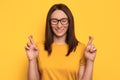 Young pretty girl with eyeglasses and crossed fingers posing on yellow background with closed eyes