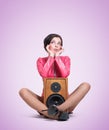Young pretty girl is sitting on the floor with a wooden loudspeaker, on a light pink background