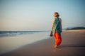 Young pretty girl walking along the seashore and admires the sunset. Blonde in bright clothes on sandy beach in the light of Royalty Free Stock Photo