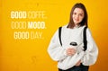 Young pretty girl in a white hoodie with a backpack and a disposable Cup of coffee poses against a yellow concrete wall.