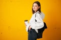 Young pretty girl in a white hoodie with a backpack and a disposable Cup of coffee poses against a yellow concrete wall.