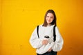 Young pretty girl in a white hoodie with a backpack and a disposable Cup of coffee poses against a yellow concrete wall.