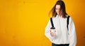 Young pretty girl in a white hoodie with a backpack and a disposable Cup of coffee poses against a yellow concrete wall.