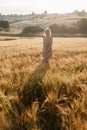 Young pretty girl in yellow dress stands at field of ears in rays of rising sun. grove and village in background Royalty Free Stock Photo