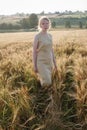 Young pretty girl in yellow dress stands at field of ears in rays of rising sun. grove and village in background