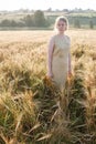 Young pretty girl in yellow dress stands at field of ears in rays of rising sun. grove and village in background Royalty Free Stock Photo