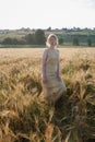 Young pretty girl in yellow dress stands at field of ears in rays of rising sun. grove and village in background Royalty Free Stock Photo