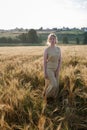 Young pretty girl in yellow dress stands at field of ears in rays of rising sun. grove and village in background Royalty Free Stock Photo