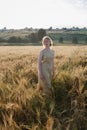 Young pretty girl in yellow dress stands at field of ears in rays of rising sun. grove and village in background Royalty Free Stock Photo