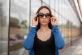 Young pretty glamorous hipster woman in a vintage knitted blue cape in fashionable sunglasses in a black t-shirt poses Royalty Free Stock Photo