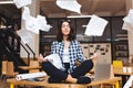 Young pretty joyful brunette woman meditating on table surround work stuff and flying papers. Cheerful mood, taking a