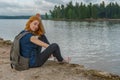 Young pretty lady with backpack sitting on bay shore in summer evening. Tourist on the beautiful landscape background. Monrepos Royalty Free Stock Photo