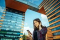 Young pretty long-haired brunette in a plaid shirt stands on city street and communicates with smartphone. Beautiful girl smiles Royalty Free Stock Photo