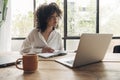 Young pretty mixed race woman studying at home with laptop. Taking notes in notebook. Bright spacious living room.