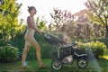Young pretty mother with baby in stroller enjoying walking in green fresh garden at sunset. Mom having fun with baby in pram in be Royalty Free Stock Photo