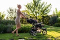 Young pretty mother with baby in stroller enjoying walking in green fresh garden at sunset. Mom having fun with baby in pram in be