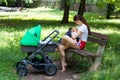 Young mother breast-feeding her cute baby, holding infant gently in hands and sitting on the park bench, next green stroller Royalty Free Stock Photo