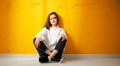 Young pretty smiling girl student in a white hoodie poses sitting on the floor against a yellow concrete wall.