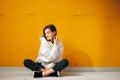 Young pretty smiling girl student in a white hoodie poses sitting on the floor against a yellow concrete wall.