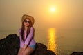 Young pretty smiling woman in straw hat, sunglasses and pink shirt sits on a rock against the sea and sky. Solar path on the water Royalty Free Stock Photo