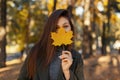 Young pretty stylish woman with blue eyes in a fashionable green coat, holding a yellow autumn leaf near the face while walking