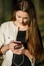 Young pretty teen girl with long hair in casual clothes surfing the internet on her smartphone in the dark forest on a sunset