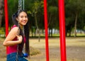 Young pretty teenage girl with pig tails wearing jeans and purple top, sitting on swing at outdoors playground, smiling Royalty Free Stock Photo