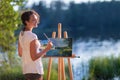 Young pretty woman artist draws paints a picture of lake on open plain air outdoors