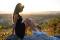 Young pretty woman in black short summer dress sitting on a rock relaxing outdoors at sunset. Fashionable female Royalty Free Stock Photo