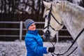 Young pretty woman in blue jacket and sports hat on a walk with a white horse on a winter cloudy day. Royalty Free Stock Photo