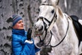 Young pretty woman in a blue jacket and a sports hat for a walk with a white horse Royalty Free Stock Photo