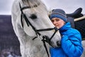 Young pretty woman in a blue jacket and a sports hat for a walk with a white horse Royalty Free Stock Photo