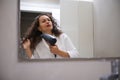 Young pretty woman drying her long beautiful curly hair with blow dryer, admiring her reflection in the bathroom mirror Royalty Free Stock Photo