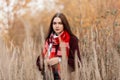 Young pretty woman with natural make-up in a stylish knitted sweater with a warm red scarf stands among tall dry grass in a field Royalty Free Stock Photo