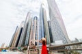 Young pretty woman in red dress and straw hat pointed with hands on downtown skycrapers in modern city. Low angle view