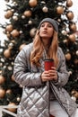 Young pretty woman with red hair in a fashionable knitted gray hat in a youth stylish silver jacket with a cup of coffee poses