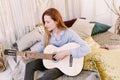 Young pretty woman with red hair studying music at home. She plays acoustic guitar and sing alone at home