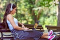 Beautiful young woman with headphones sitting on bench in the park, using laptop computer Royalty Free Stock Photo