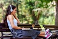 Beautiful young woman with headphones sitting on bench in the park, using laptop computer Royalty Free Stock Photo