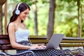 Young pretty woman with headphones sitting on bench in the park, using laptop computer Royalty Free Stock Photo