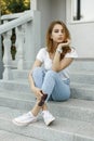 Young pretty woman in stylish T-shirt in jeans in trendy sneakers is resting sitting on the steps of a vintage building. Royalty Free Stock Photo