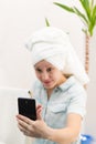 A young pretty woman with a towel on her head in a bathroom with a toothbrush and a smartphone in her hand brushes her teeth