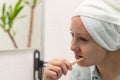 A young pretty woman with a towel on her head in a bright bathroom with a toothbrush in her hand brushes her teeth. Reflection in