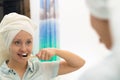 A young pretty woman with a towel on her head in a bright bathroom with a toothbrush in her hand brushes her teeth. Reflection in