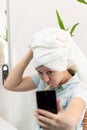 A young pretty woman with a towel on her head in a bright bathroom with a toothbrush and a smartphone