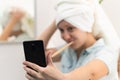 A young pretty woman with a towel on her head in a bright bathroom with a toothbrush and a smartphone in her hand brushes her teet