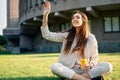 Young pretty woman welcome friends waving raised palm, sitting on grass with coffee