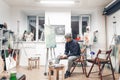 Young professional artist sits on a chair near an easel with a host in a cozy creative studio and paints an oil painting. Creative