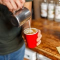 Young professional barista pours cream from a metal cup into a hot delicious latte. Man makes coffee. Close-up.